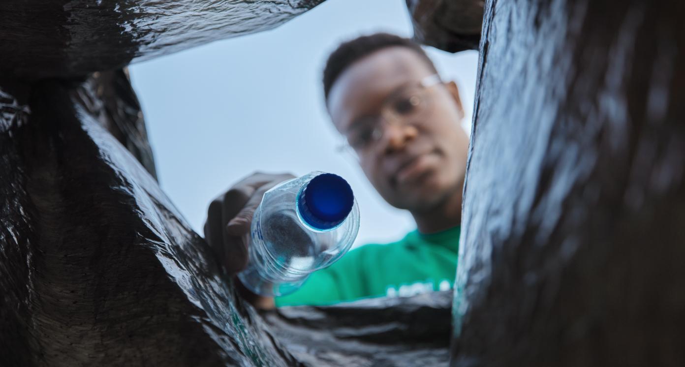 Recycle, bottle and black man with view in bag, sustainability and cleaning plastic pollution, earth day and community service. Saving the environment, charity and people putting trash in garbage bin